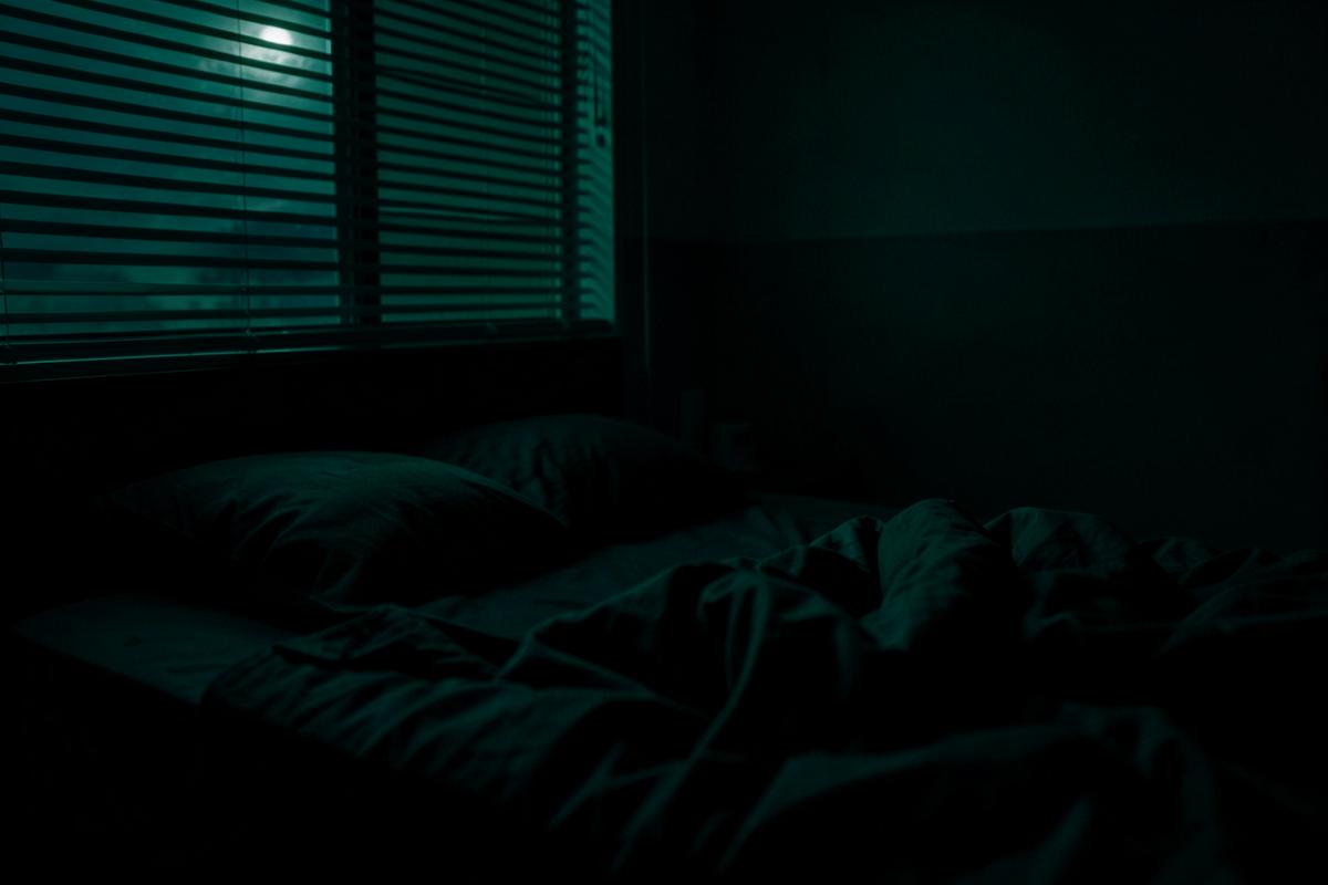 Empty bed with rumpled dark bedding in a quiet bedroom lit by faint moonlight through blinds